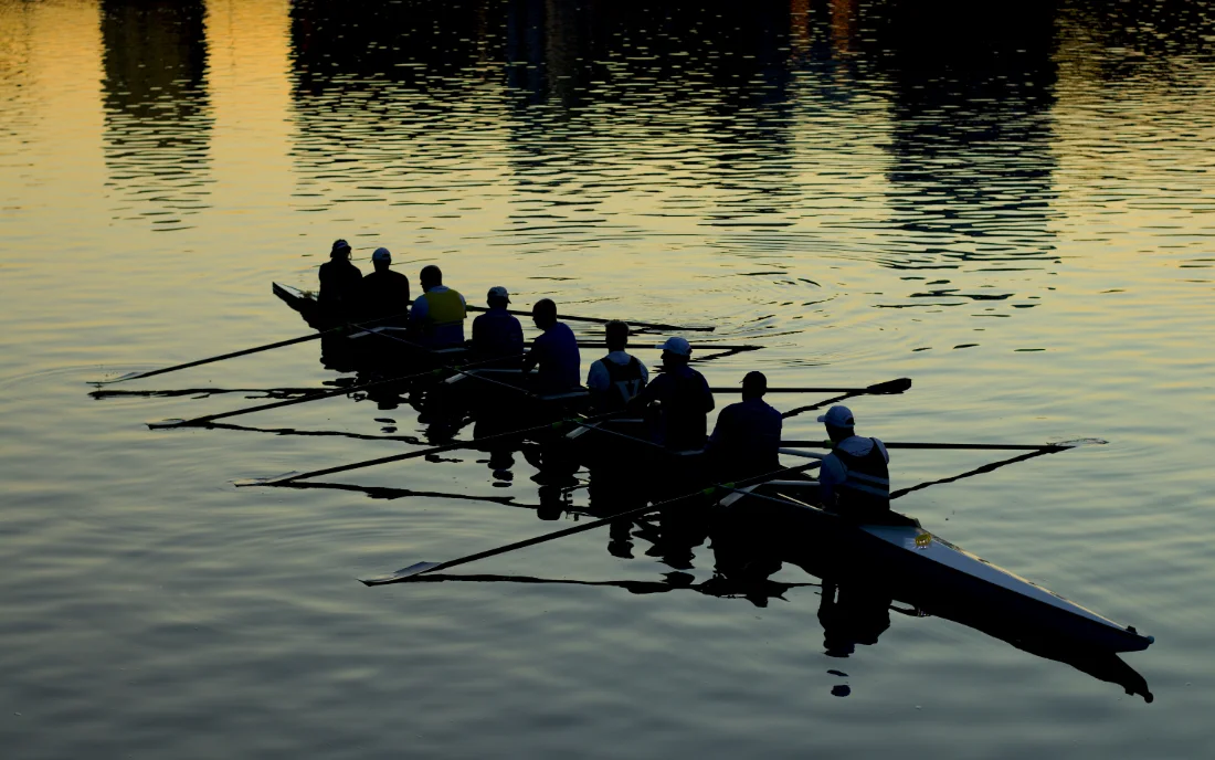 A rowing eight moving across still water at dusk, every oar at the same angle.