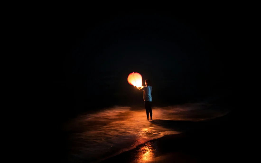 One figure alone on a wet beach at night, holding up a lit lantern.