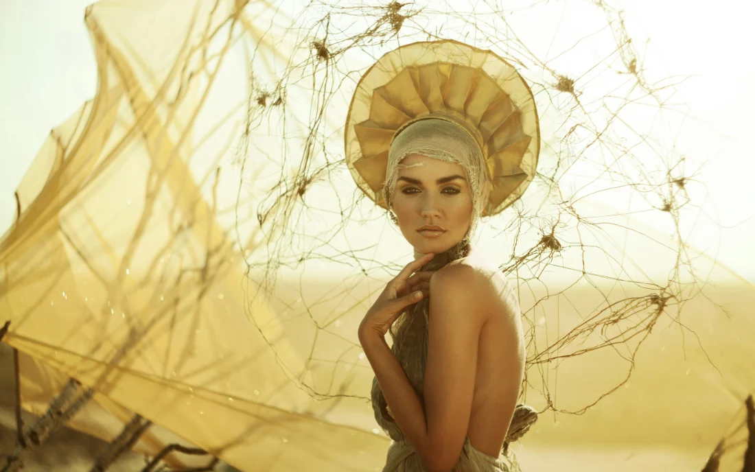 A woman in a gold headdress backlit in a desert, fine netting lifting in the wind.