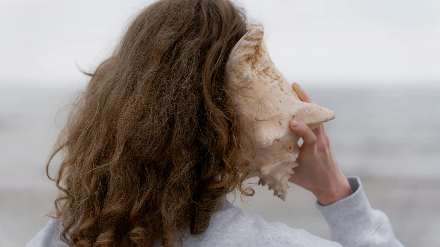 A person on a beach holding a large conch shell to their ear.