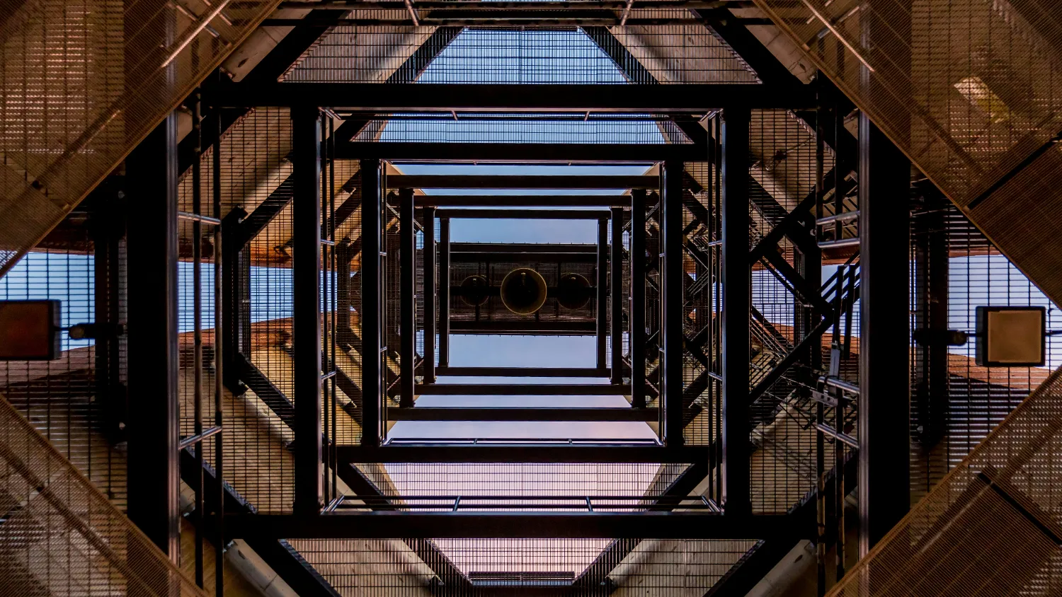 Looking up through a repeating steel structure towards the sky.