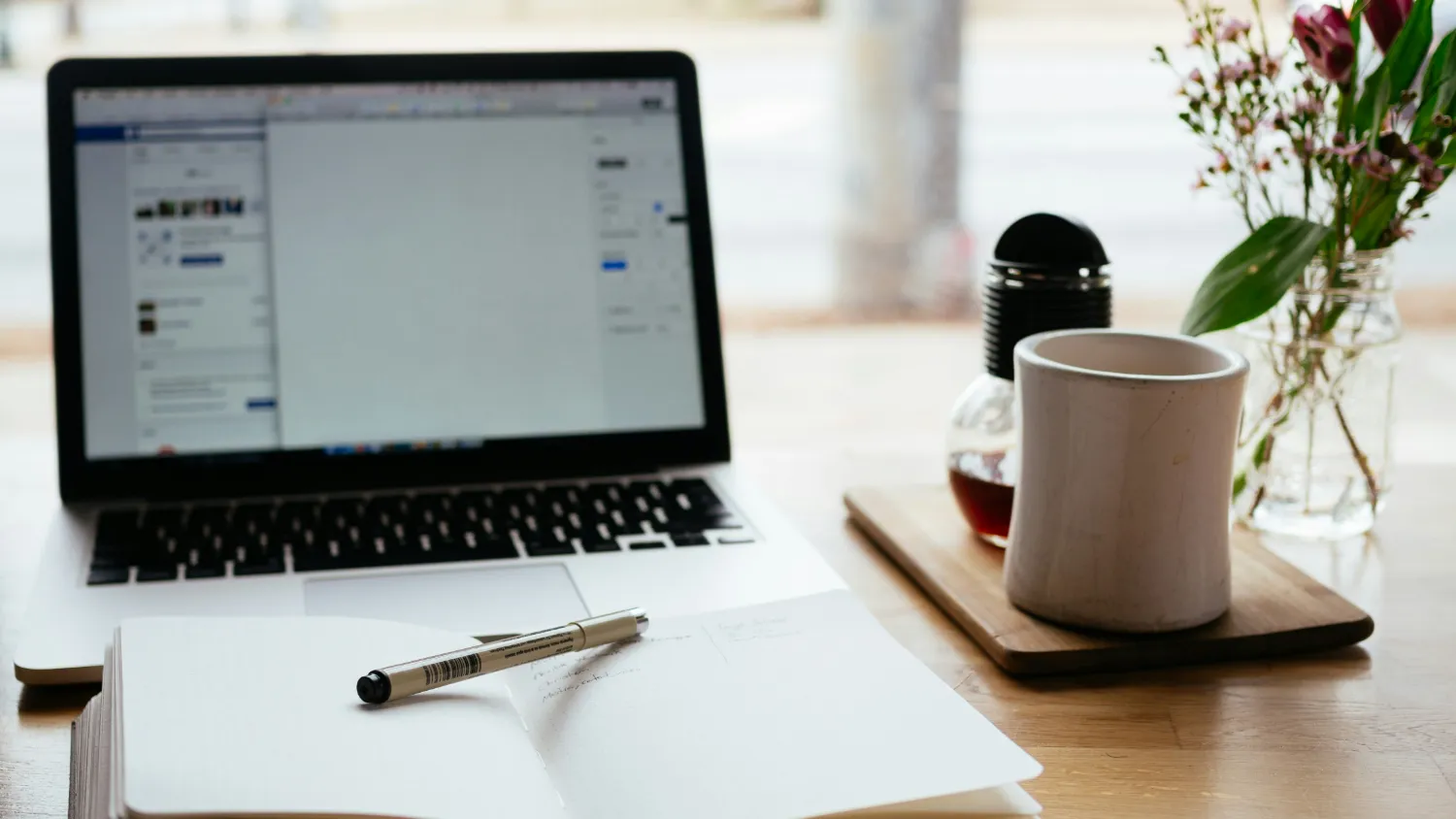 A laptop, a notepad and a pen on a desk beside a cup of coffee.