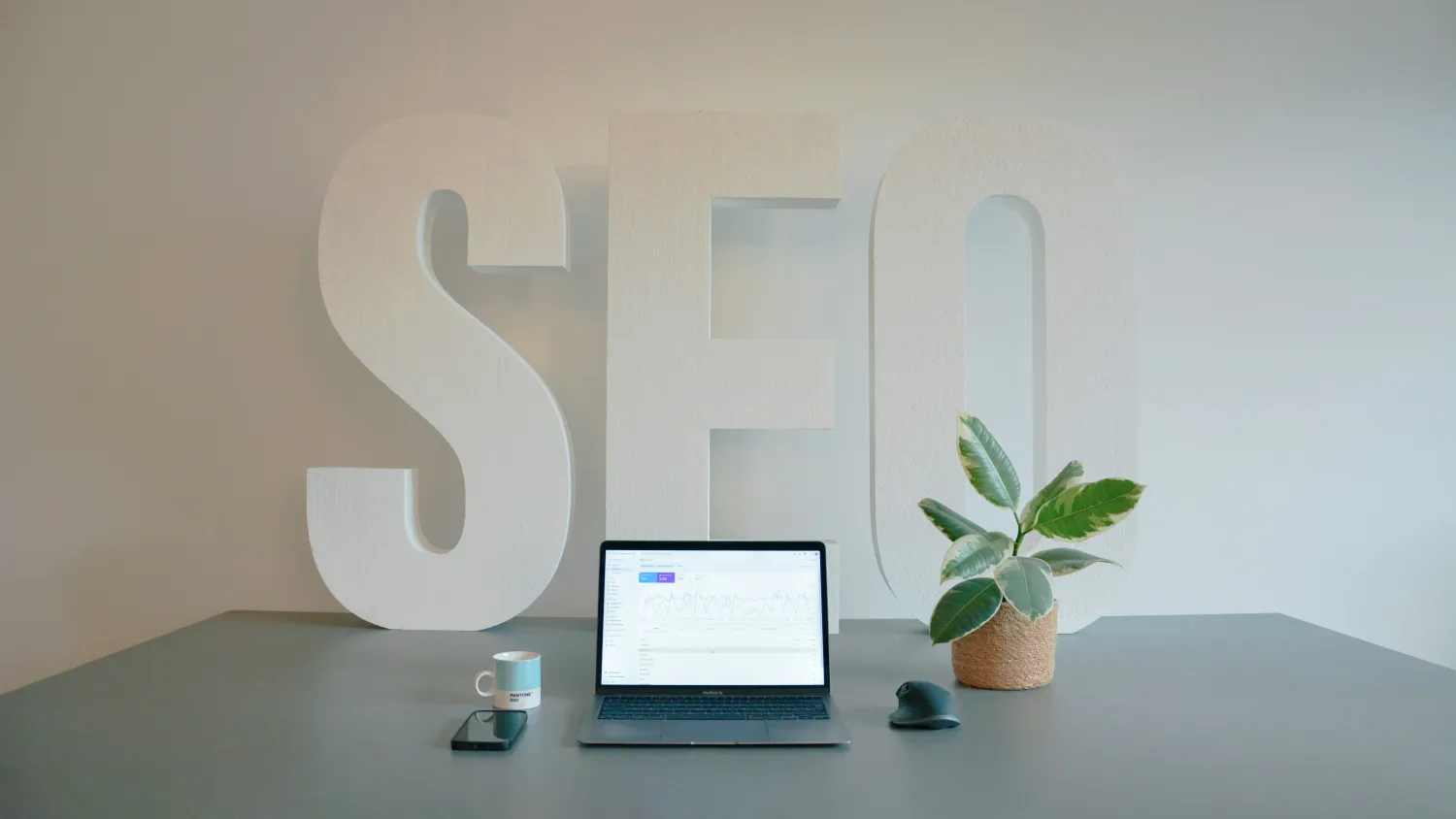The letters S E O standing on a desk behind an open laptop and a plant.