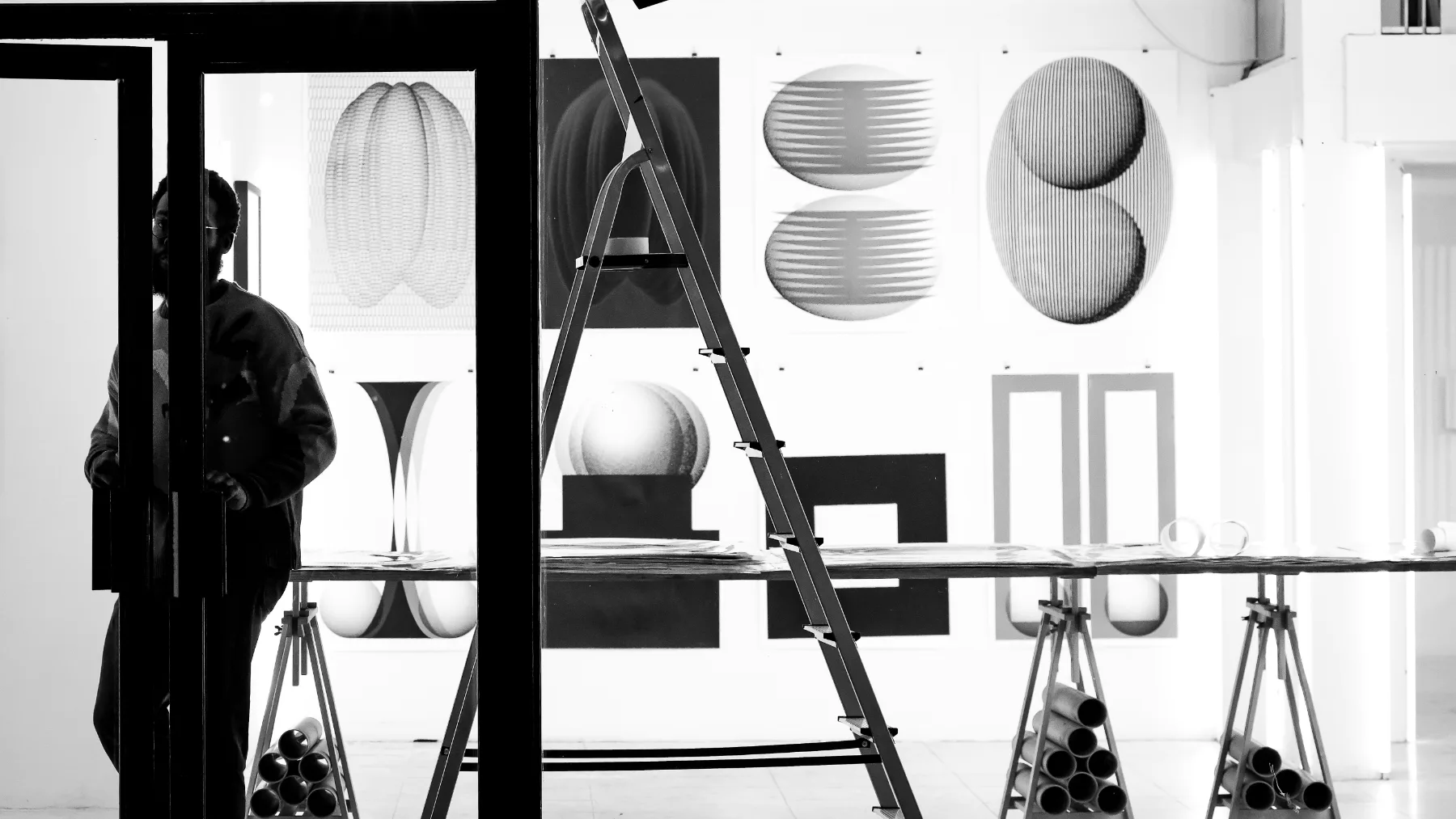 A studio wall hung with printed work in progress, one person at the door.